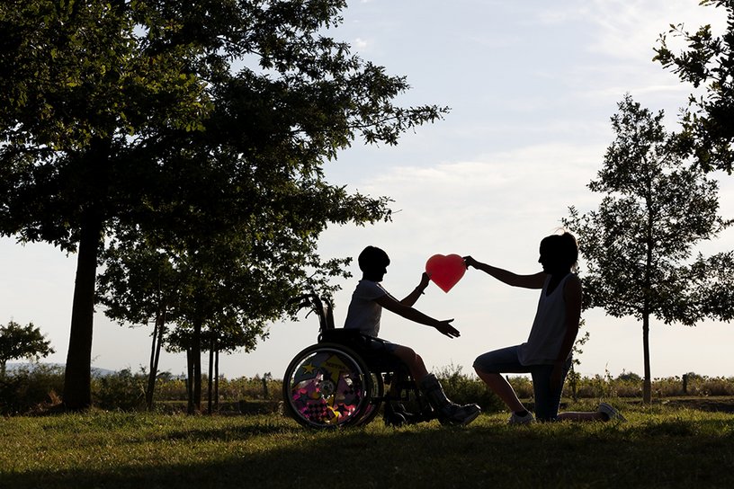 Ein Kind im Rollstuhl hält gemeinsam mit einer jungen Frau ein Herz hoch.