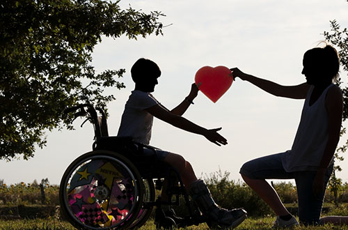Ein Kind im Rollstuhl und eine Frau halten ein rotes Herz gemeinsam hoch.